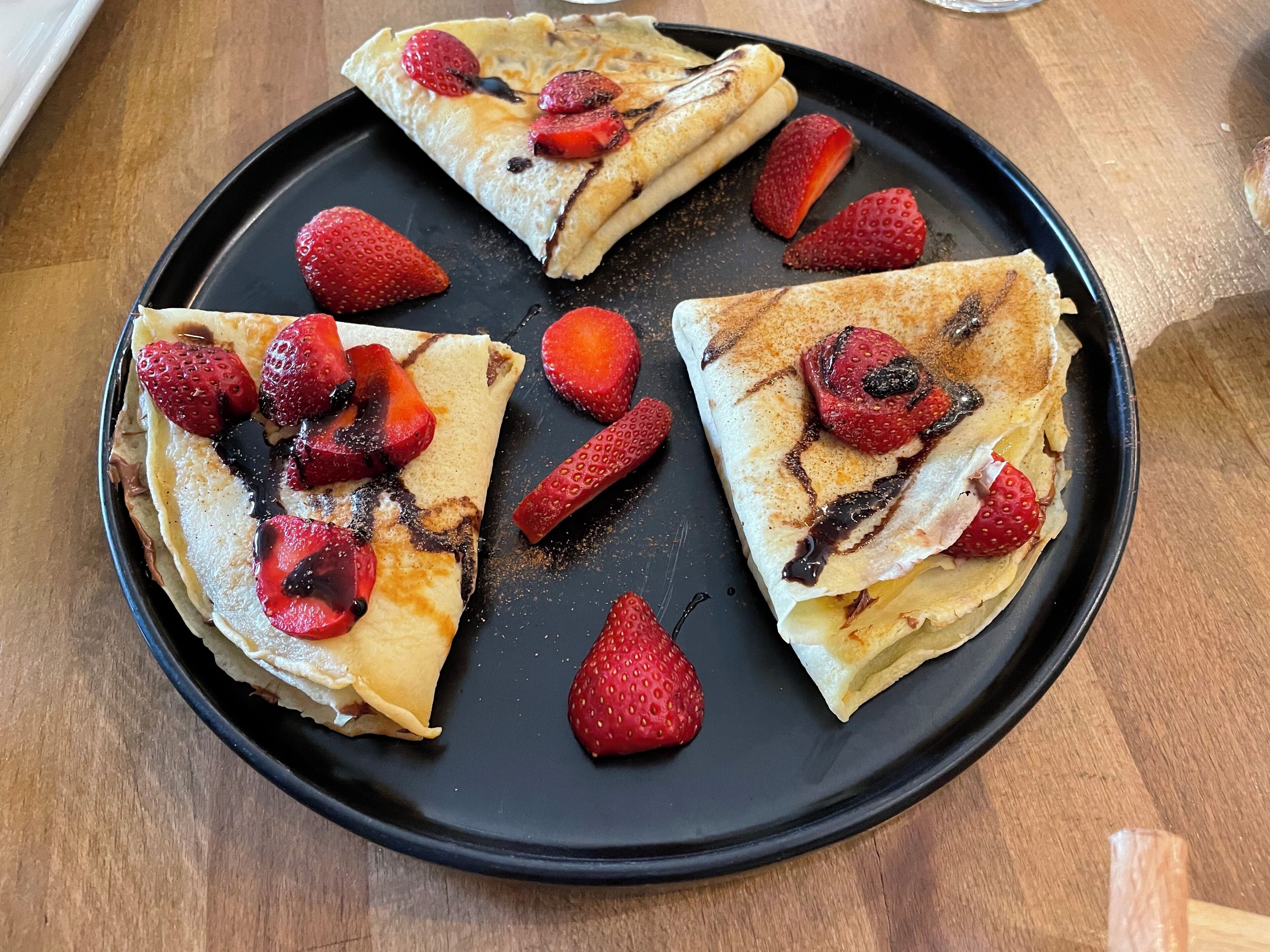 Une assiette noire sur laquelle sont disposées trois crêpes, garnies de fraises et nappées de sauce au chocolat.