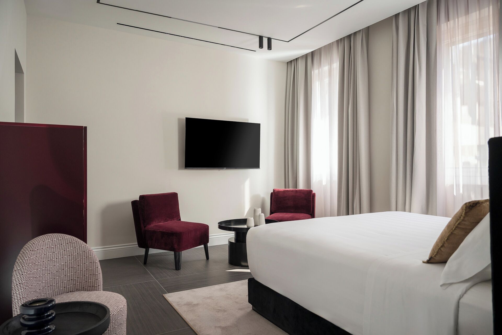 A modern hotel room with a large bed, two red chairs, a small table, and a flat-screen TV mounted on the wall.