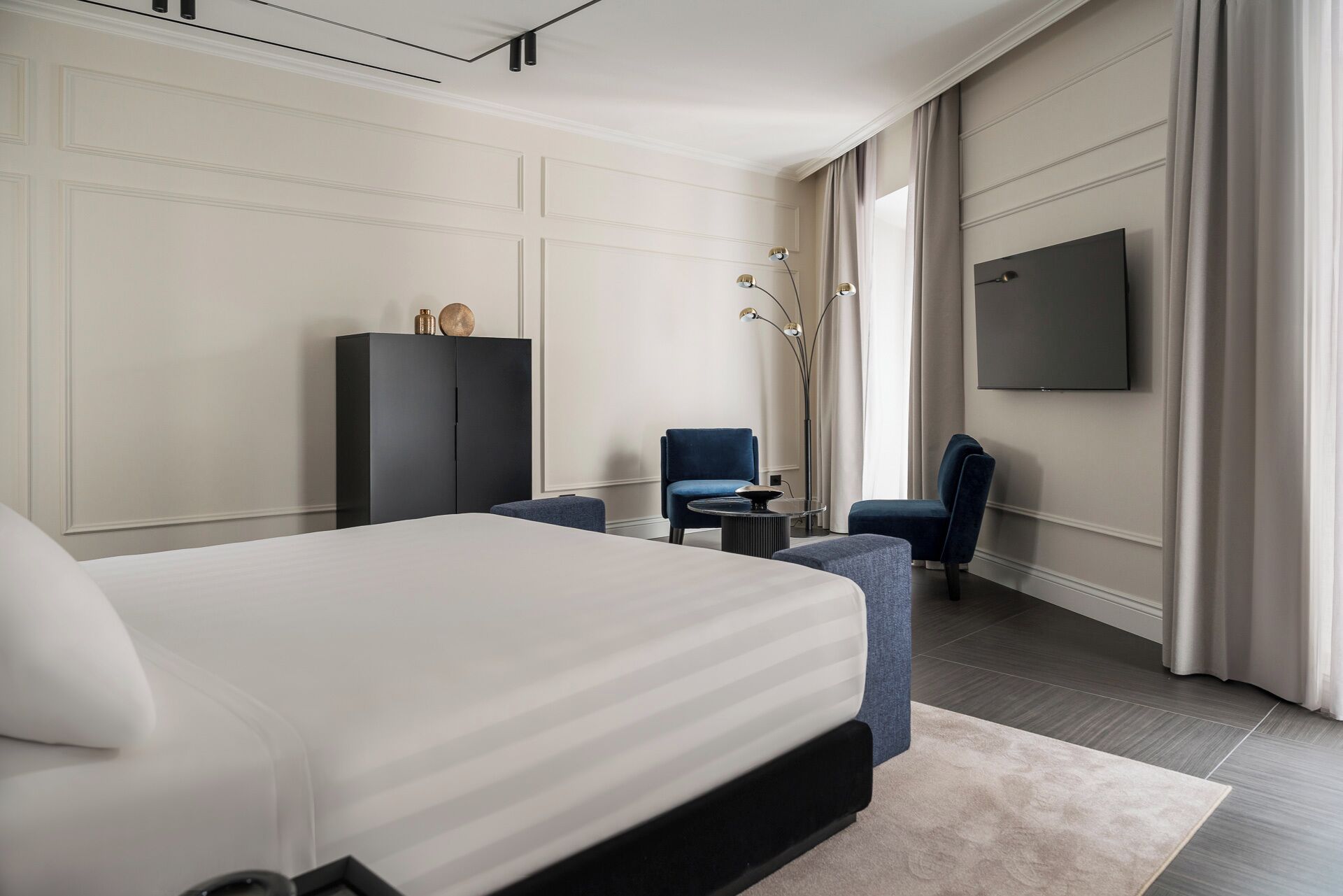 A modern hotel room with a large bed, a desk, two chairs, a TV, and a wardrobe.