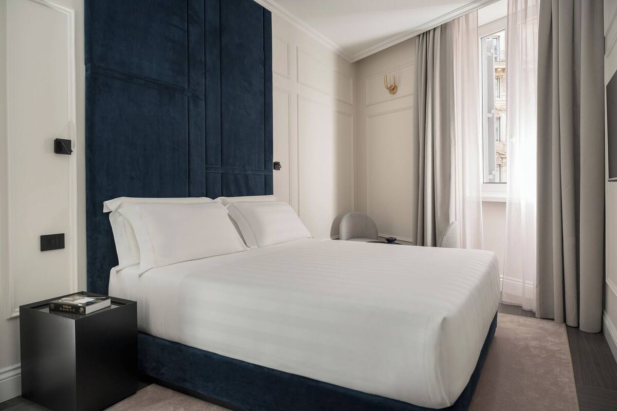A neatly made bed in a hotel room with a dark blue headboard, a small bedside table with a book, and a chair.