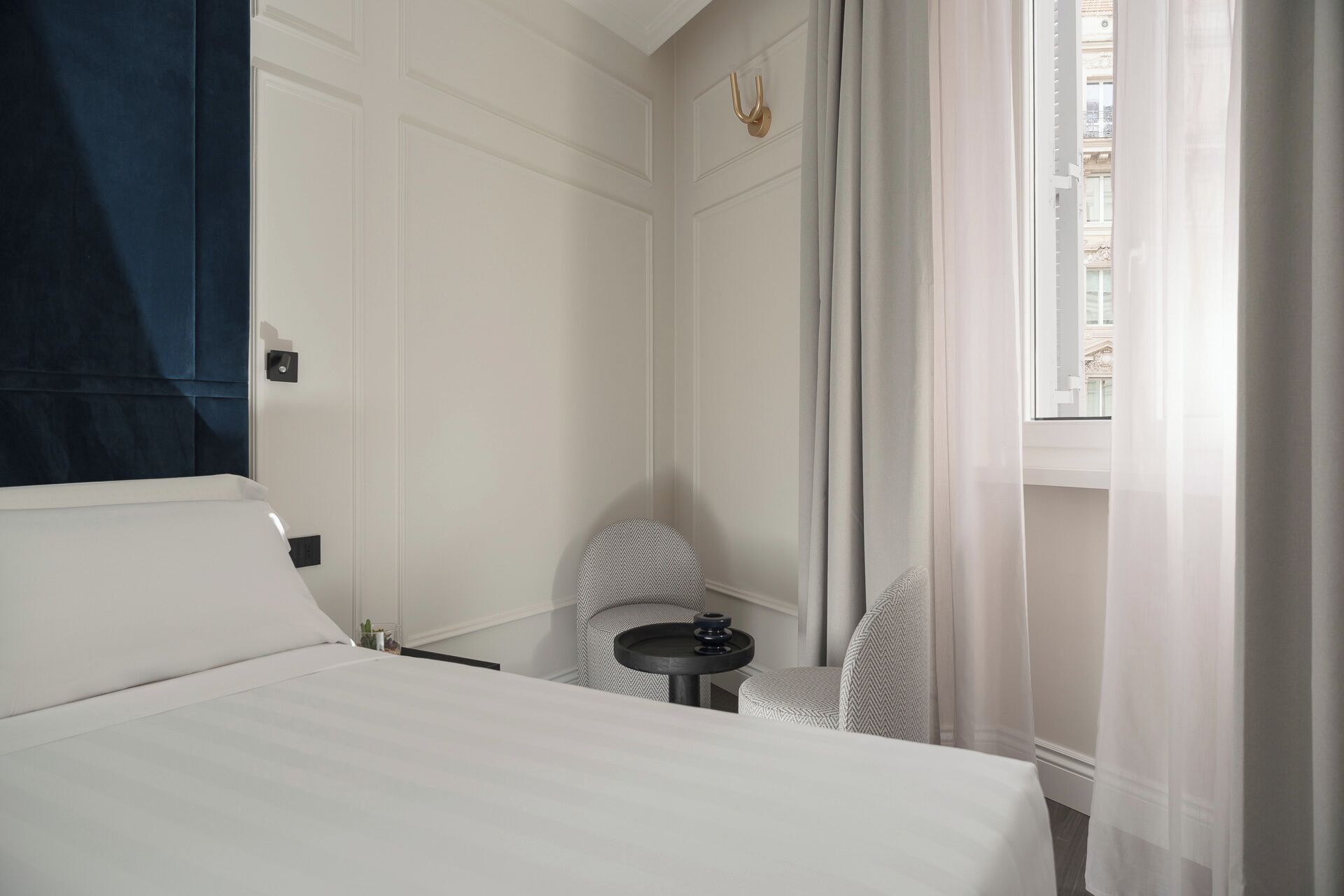A hotel room with a bed, a small table, a chair, and a window with curtains.