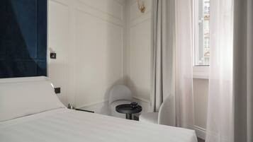 A hotel room with a bed, a small table, a chair, and a window with curtains.