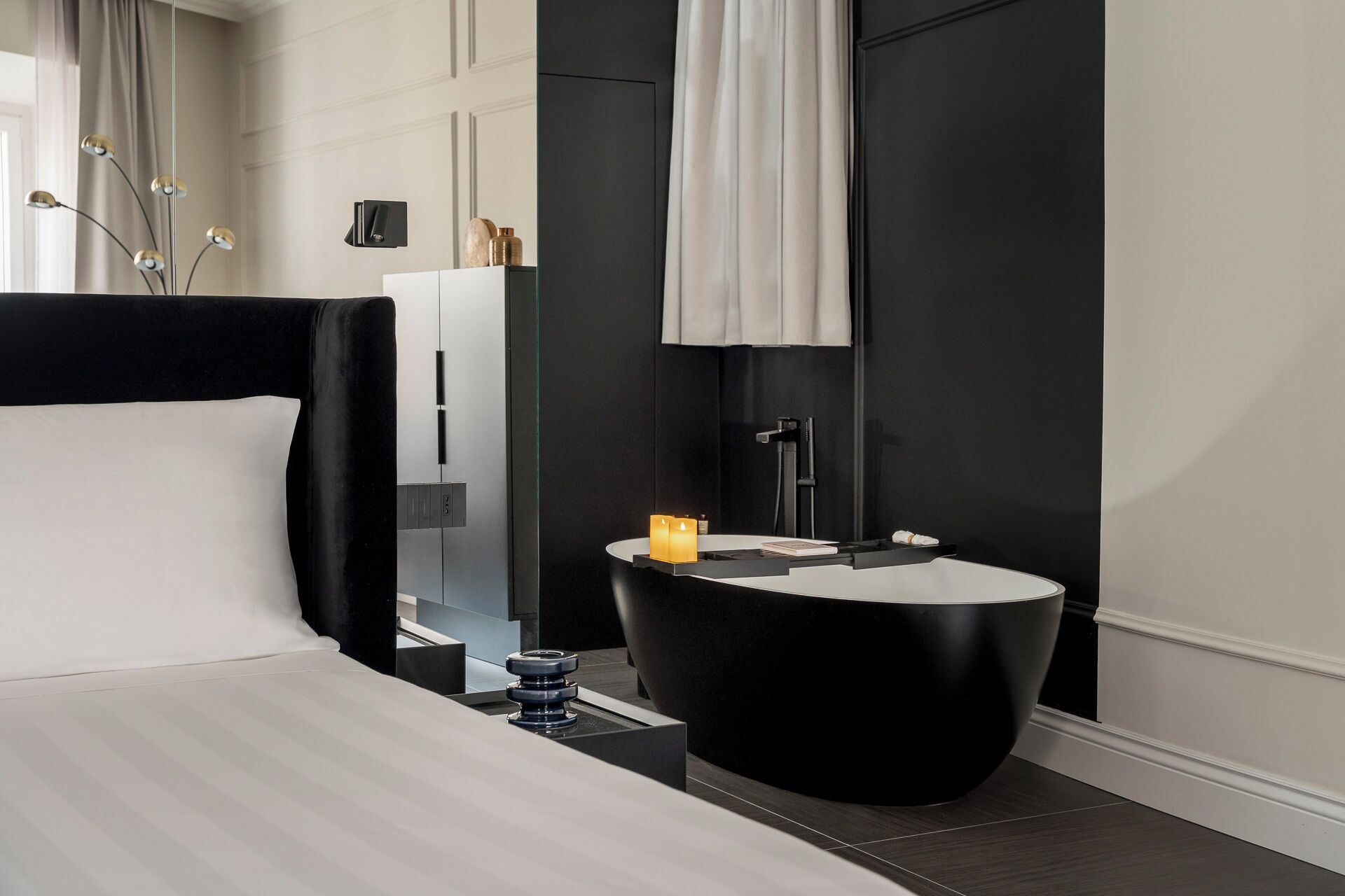 A modern hotel room with a black and white color scheme, featuring a large bed, a round black side table with a candle, a black bathtub, and a large mirror.