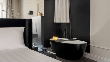 A modern hotel room with a black and white color scheme, featuring a large bed, a round black side table with a candle, a black bathtub, and a large mirror.