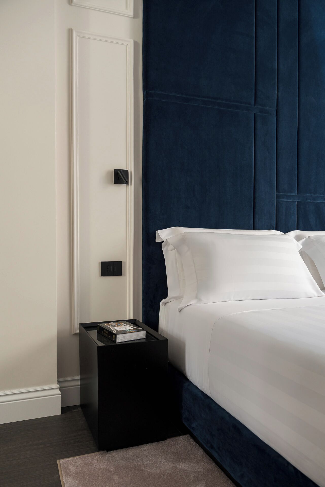 Un dormitorio moderno con un cabecero azul oscuro, ropa de cama blanca y una mesita de noche negra con un libro.