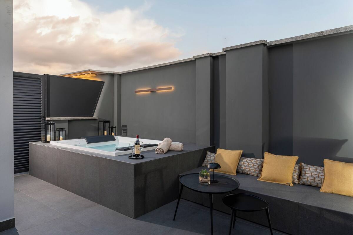A modern outdoor seating area with a built-in hot tub, a small table with a lamp, and a sofa with cushions.