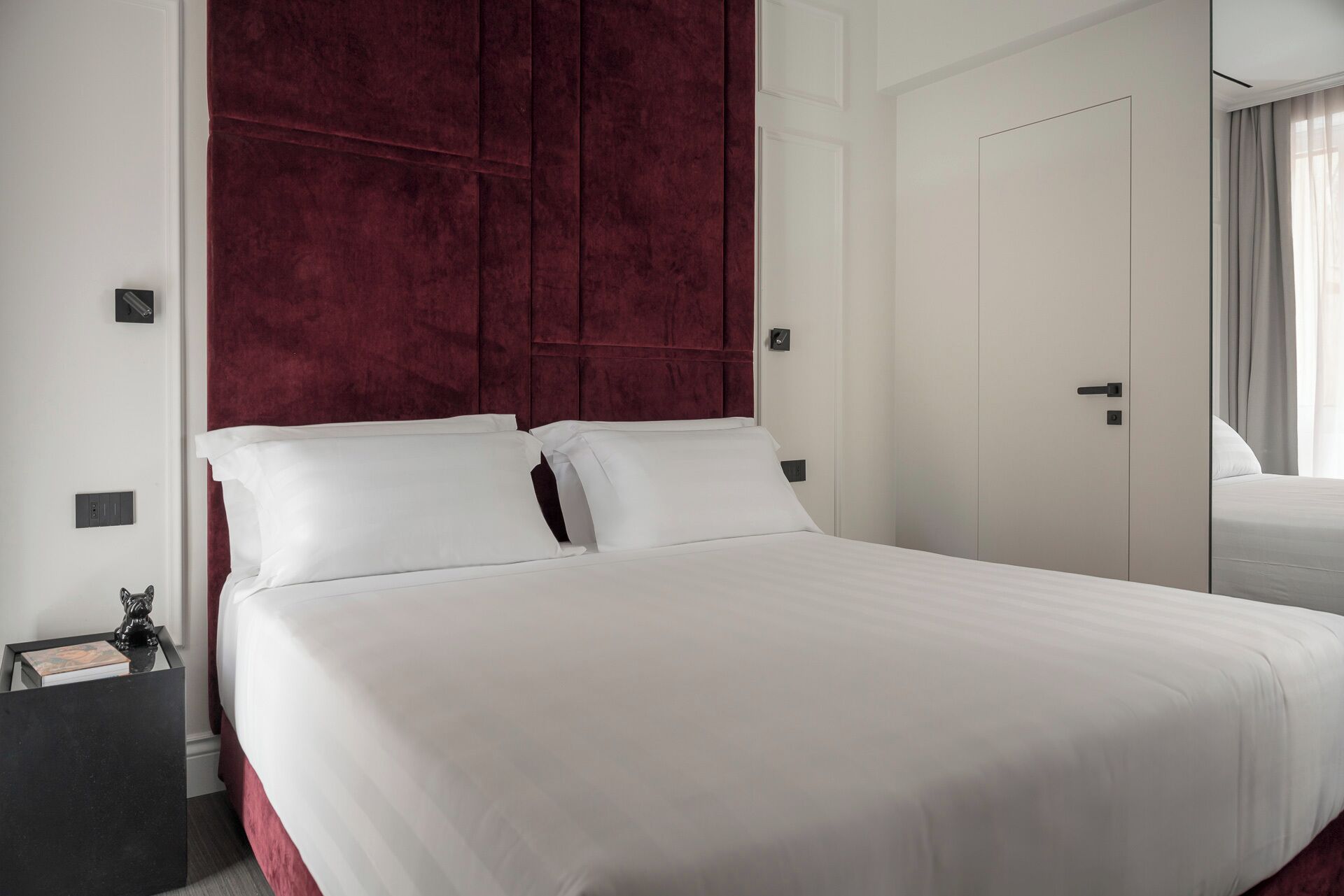 A hotel room with a large bed, a red headboard, a nightstand, and a door with a handle.