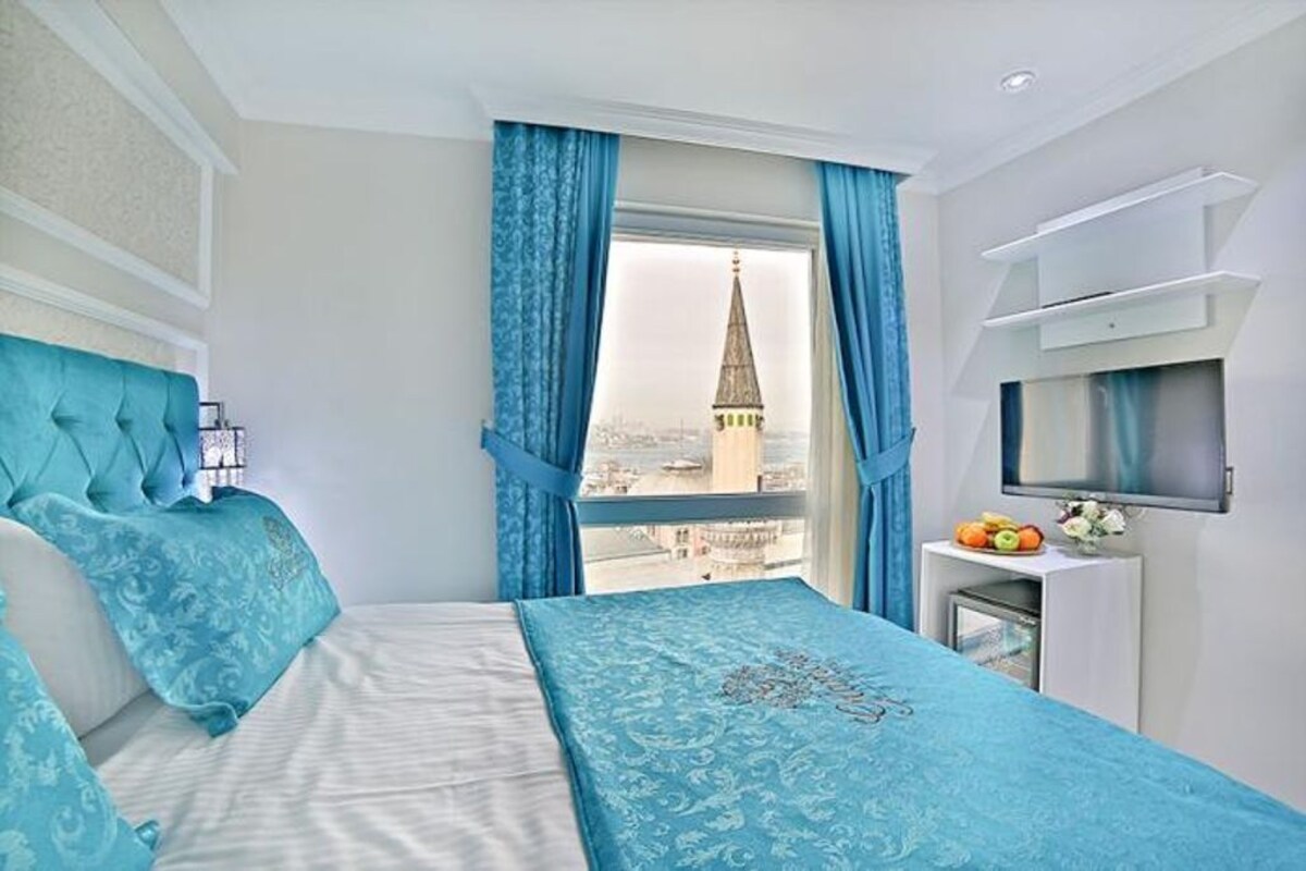 Habitación de hotel con cama, televisión, pequeña mesa con fruta y ventana con cortinas azules.