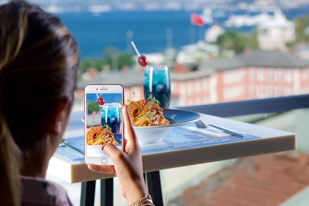 Eine Person hält ein Smartphone mit einem Foto einer Mahlzeit auf einem Balkon mit Blick auf ein Gewässer und Gebäude.