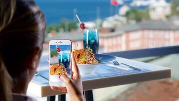 Eine Person hält ein Smartphone mit einem Foto einer Mahlzeit auf einem Balkon mit Blick auf ein Gewässer und Gebäude.