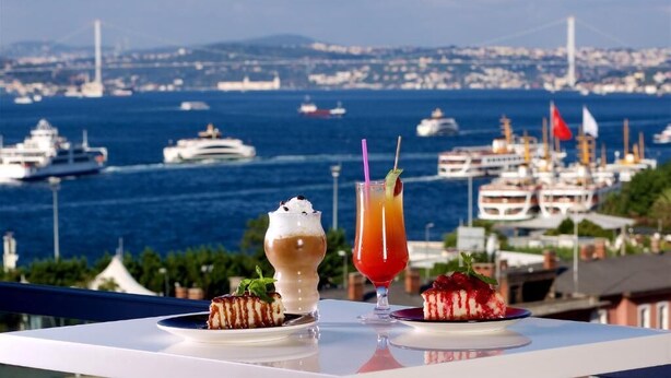 A table with two desserts and two drinks, overlooking a marina with boats and a bridge. - Glamour Hotel Istanbul Sirkeci (Istanbul)