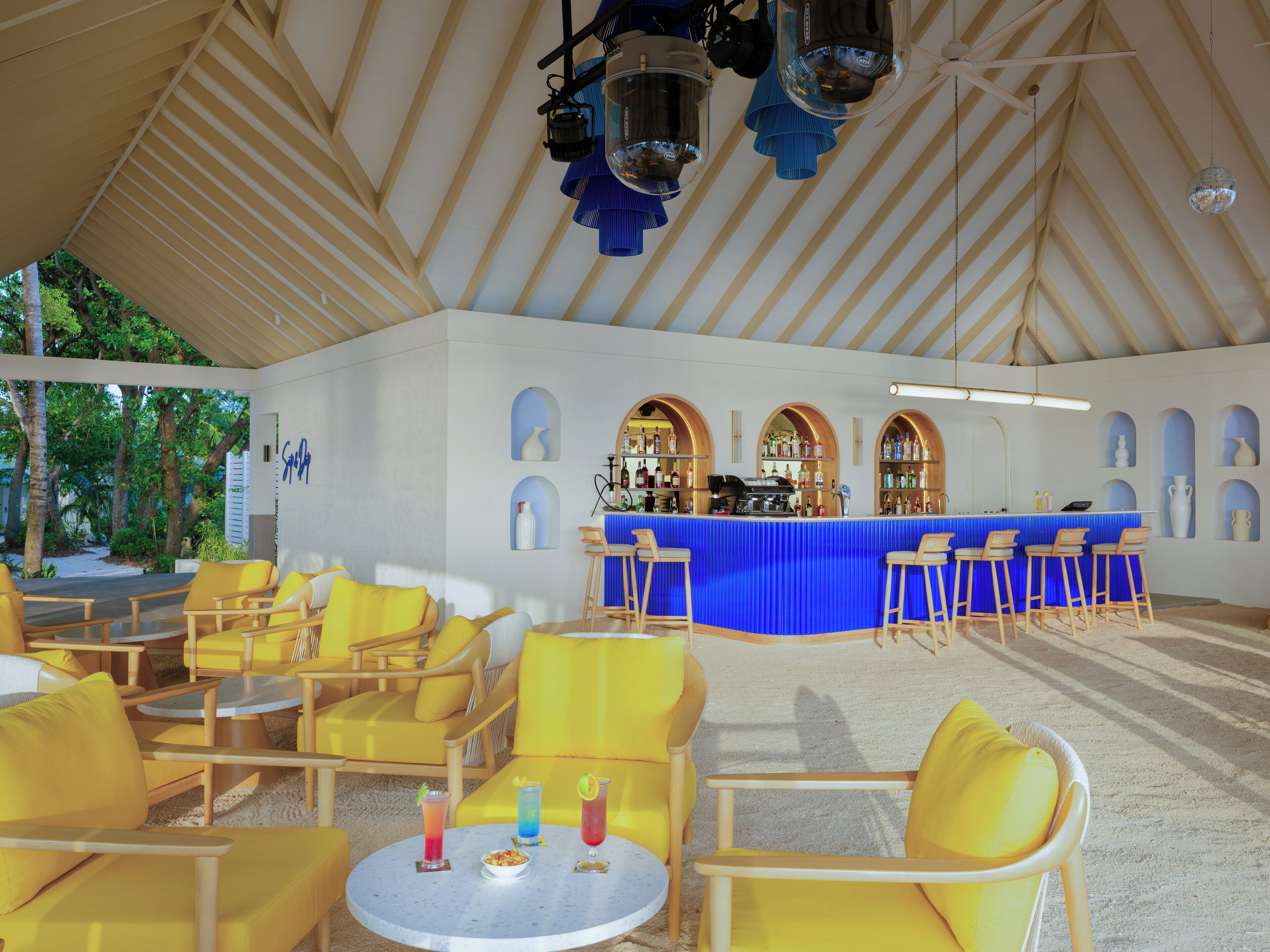 Un espace extérieur aménagé avec des sièges, protégé par un toit, comprenant des chaises et des tables jaunes, un bar aux touches de bleu et des fenêtres en arc de cercle.