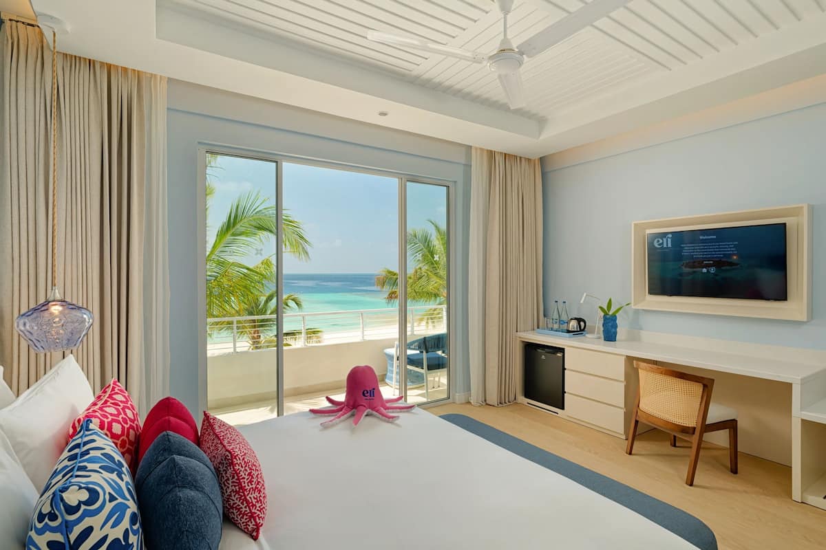 A hotel room with a large bed, a desk, a TV, and a balcony with a view of the beach.