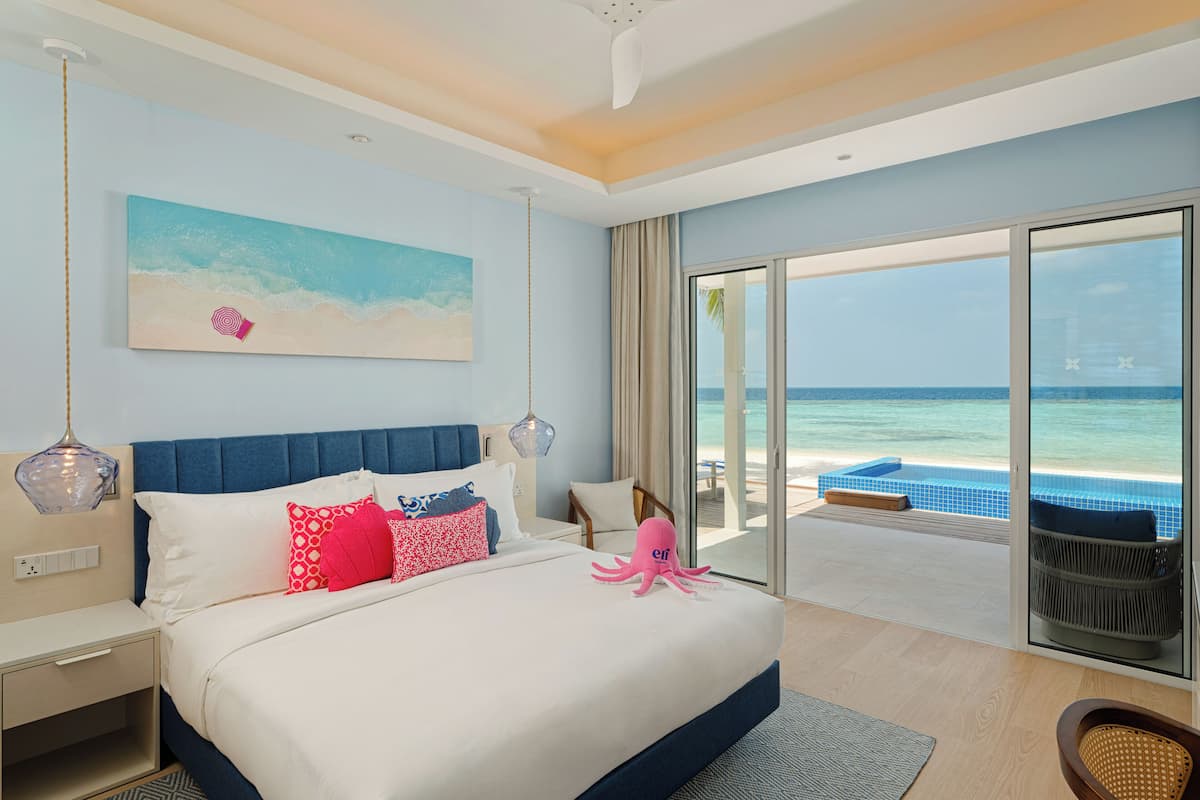 A hotel room with a bed, a view of the ocean, a beach, and a pool.