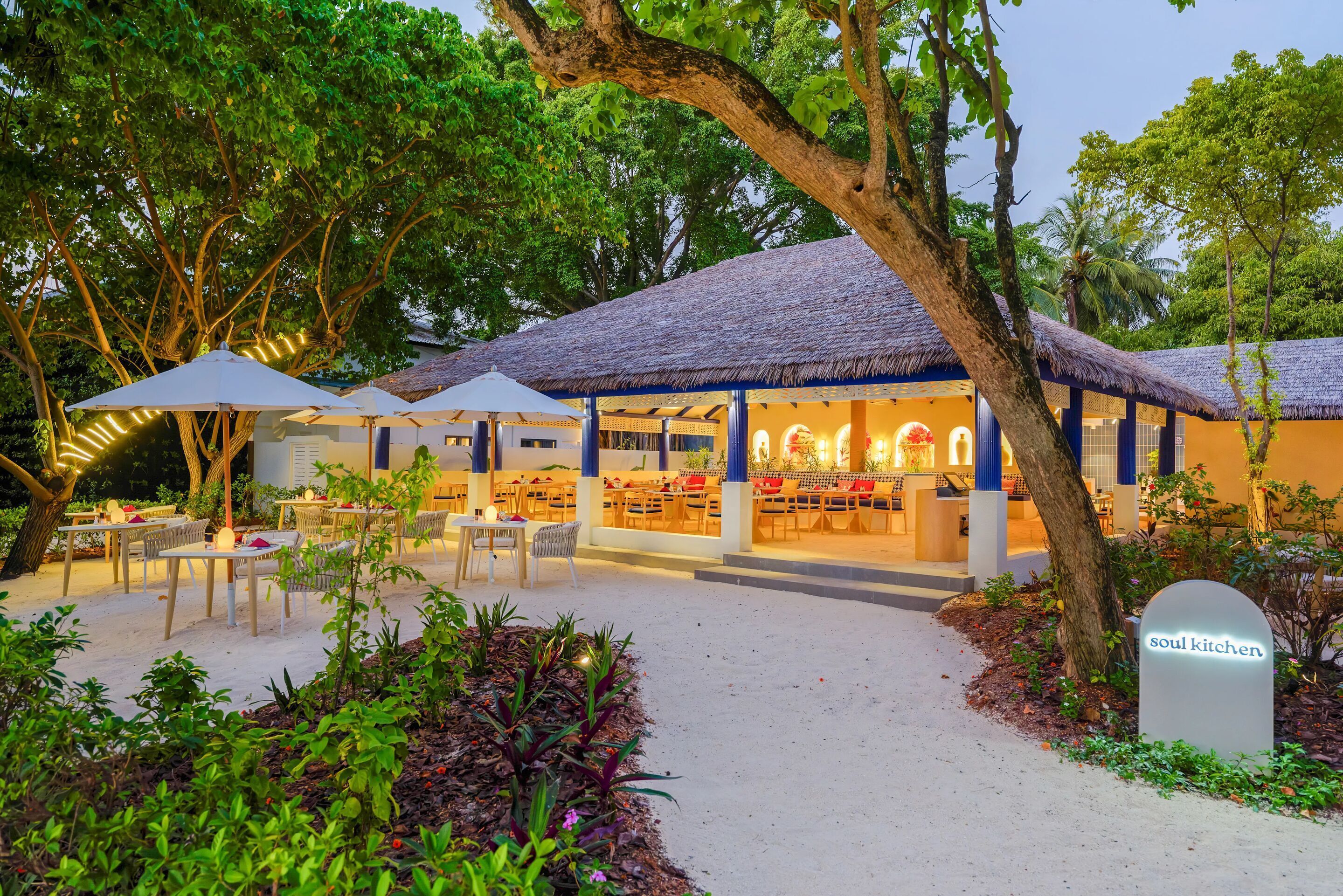 An outdoor dining area with tables and umbrellas, surrounded by lush greenery and trees.