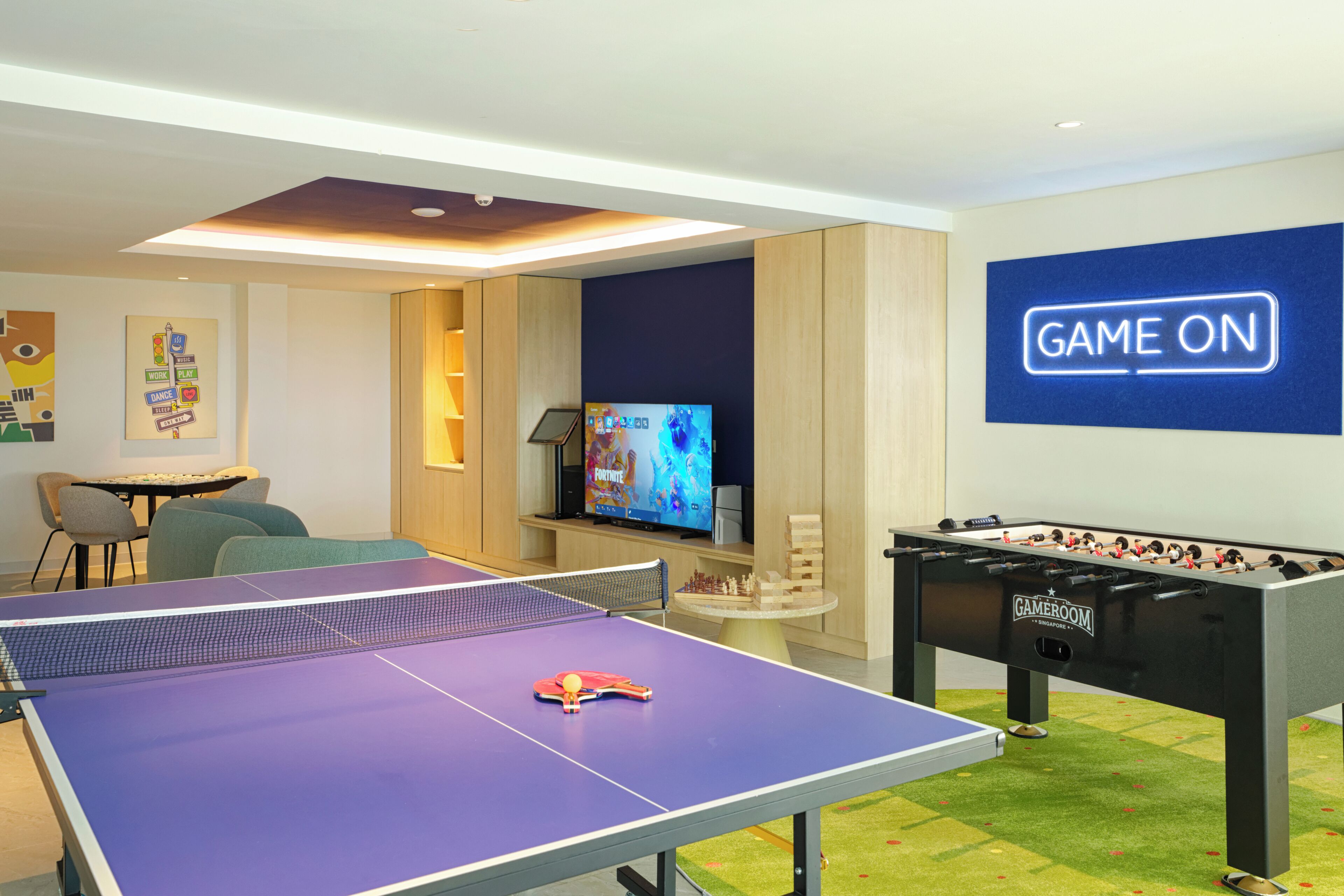 Um quarto com mesa de pingue-pongue, mesa de futebol de mesa, televisão e um letreiro com a inscrição 'GAME ON'.