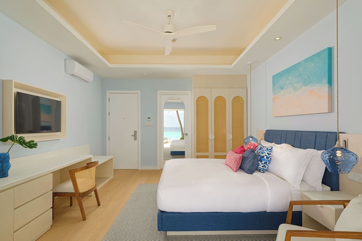 A bedroom with a bed, a desk, a chair, a TV, and a view of the beach through a door.