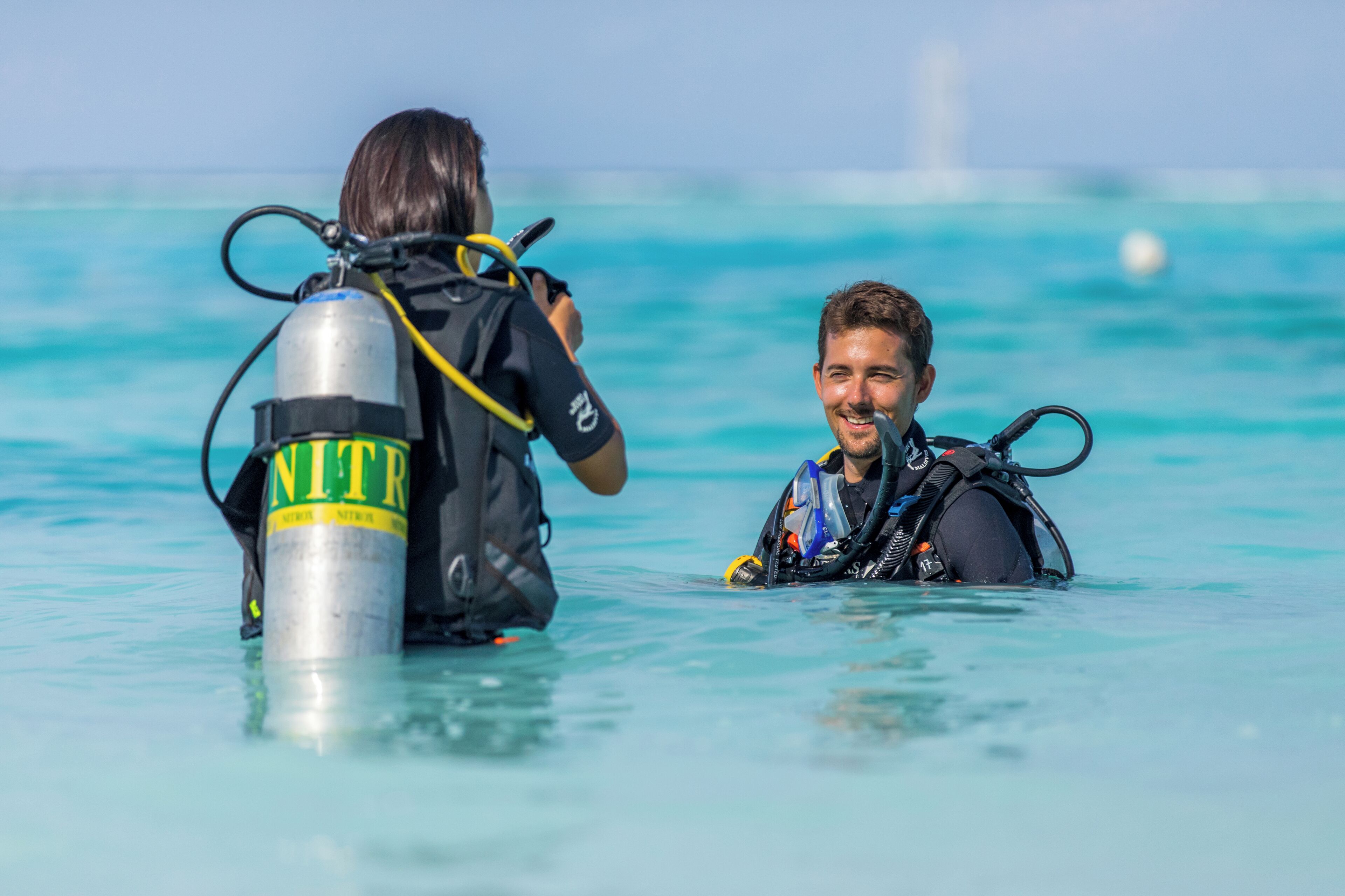 Dos personas en un mar azul transparente, una sosteniendo un tanque de oxígeno grande.
