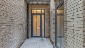 A narrow outdoor corridor with a wooden floor, a glass door with a dark frame, and a brick wall on one side. - Wimpole Mews - 4 Bedroom Apartment (London)