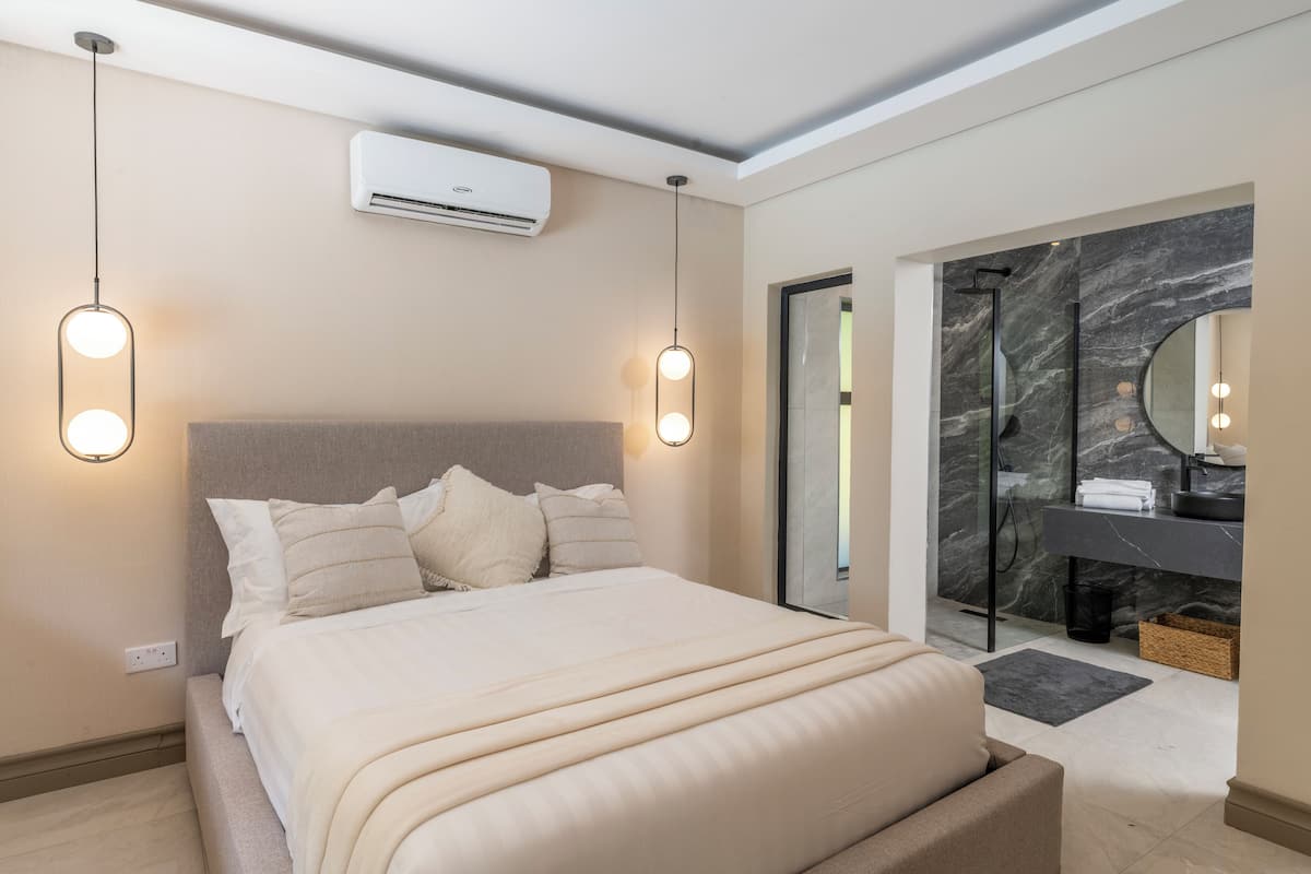 Un dormitorio moderno con una cama grande, lámparas de noche, aire acondicionado y un baño con pared y lavamanos de mármol.