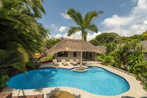 Une piscine dotée d’une structure à toit de chaume, de chaises longues et d’une végétation tropicale.