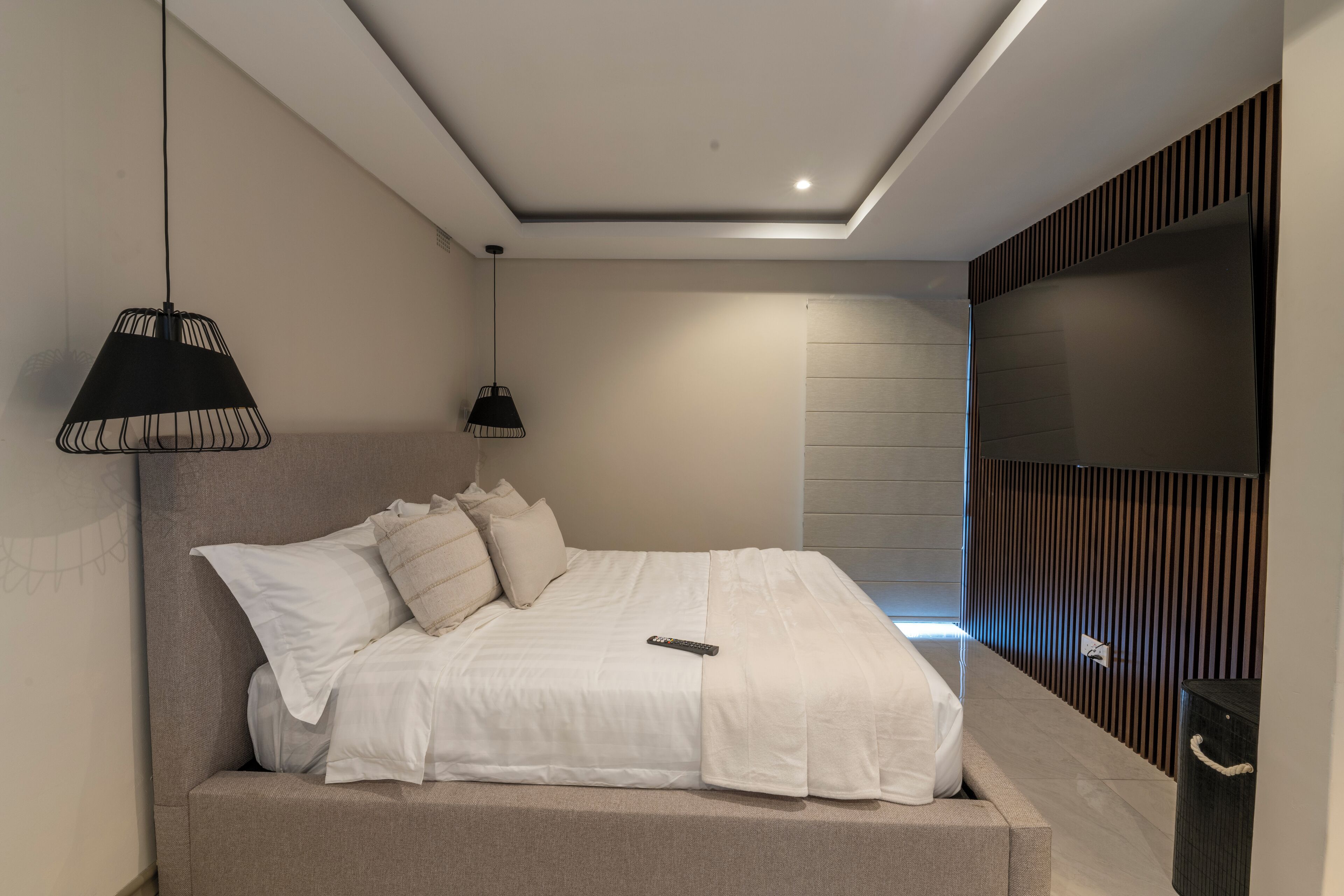 Une chambre moderne avec un grand lit, deux lampes suspendues, un téléviseur fixé au mur et un décor mural en panneaux de bois.