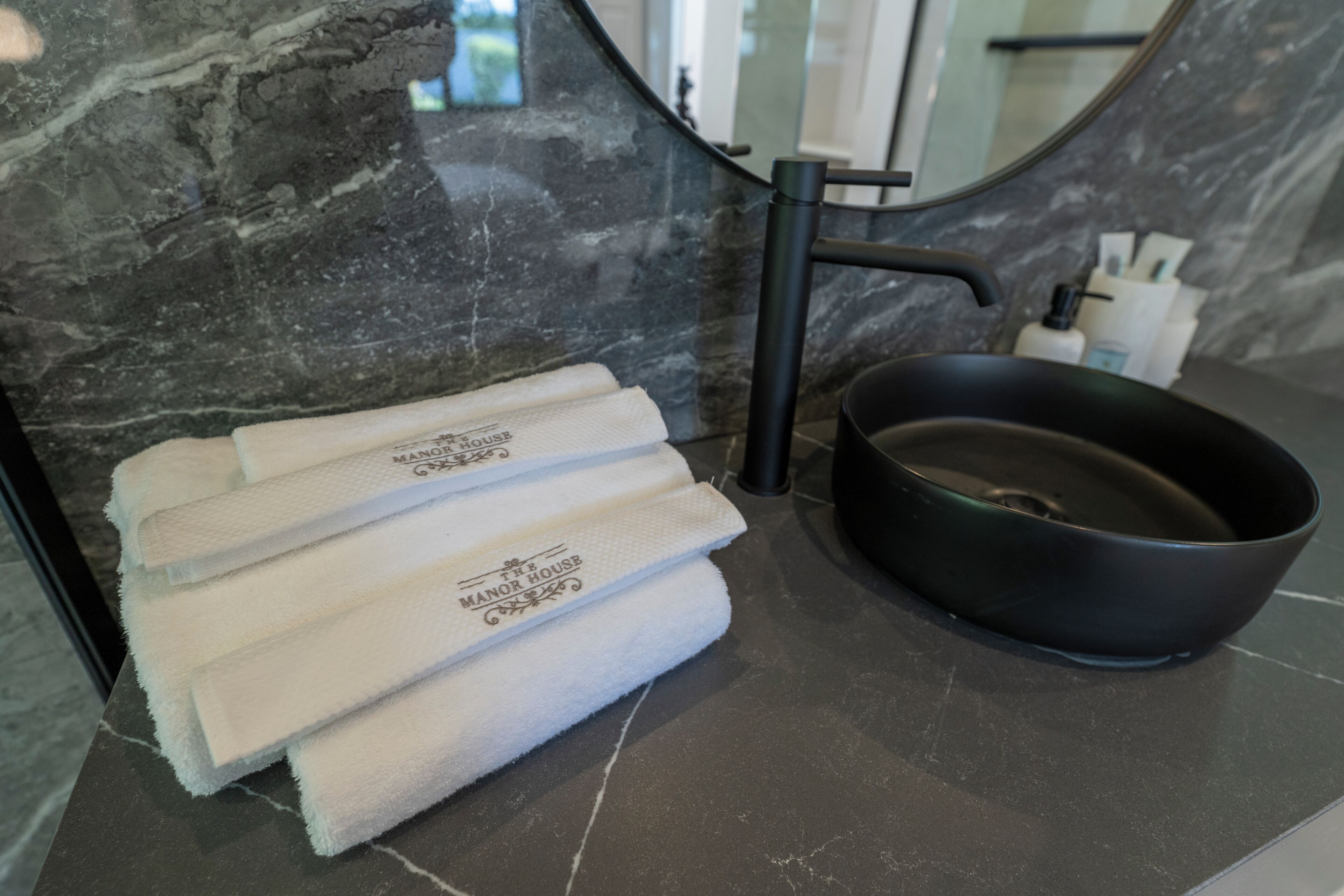 A bathroom with a marble countertop, a black sink, and a bowl. There are white towels stacked on the countertop with a logo.