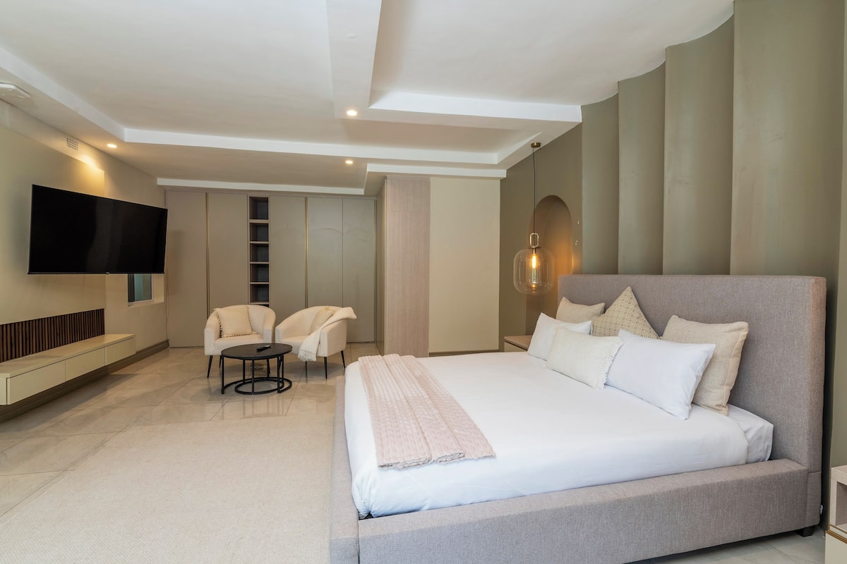 Un dormitorio moderno con una cama grande, un televisor de pantalla plana, una zona de estar con sillas y un armario empotrado.