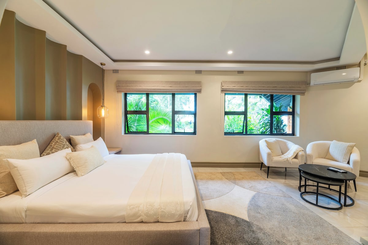 Un dormitorio moderno con una cama grande, dos sillones, una mesita y ventanas amplias con vistas a la vegetación.