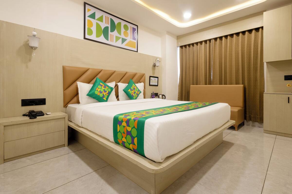 A hotel room with a large bed, a nightstand, a wall-mounted lamp, a framed picture, and a curtain.