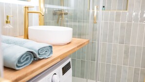 A modern bathroom with a washing machine, a sink with a gold faucet, and a glass shower enclosure. - The Royal Hues (Reims)
