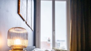 A bed with a view of a cityscape through a window, a bedside table with a lamp, and a wall with framed pictures. - The Royal Hues (Reims)