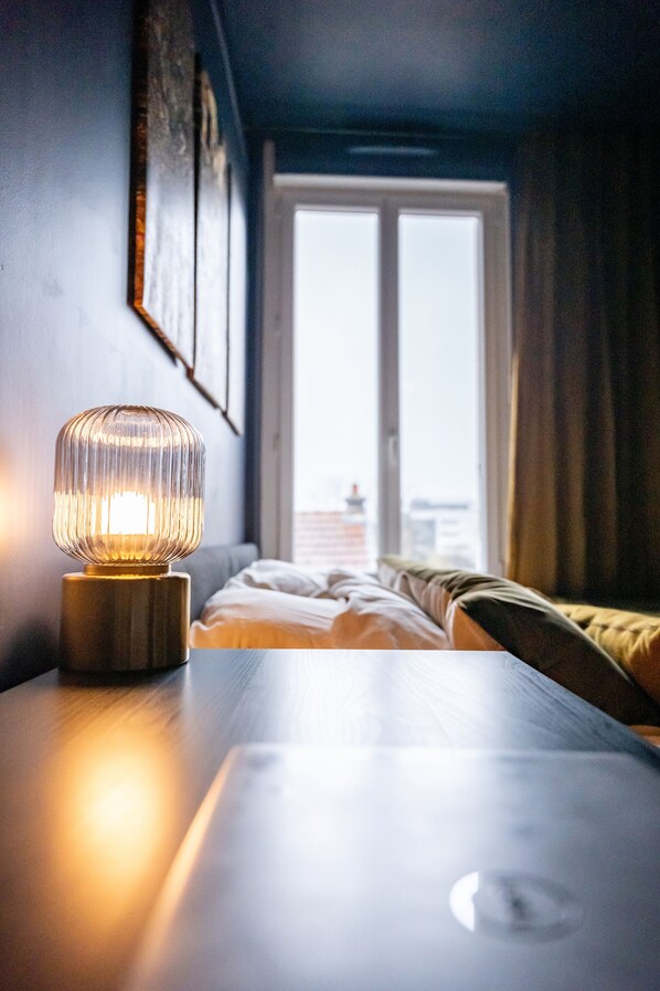 A bed with a view of a cityscape through a window, a bedside table with a lamp, and a wall with framed pictures.