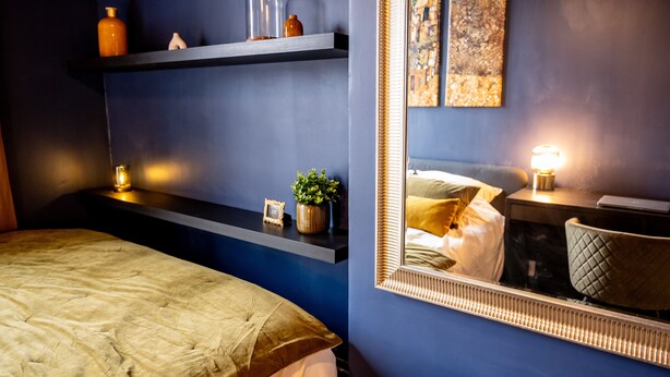 A bedroom with a bed, a bedside table with a lamp, a shelf with decorative items, and a large mirror. - The Royal Hues (Reims)