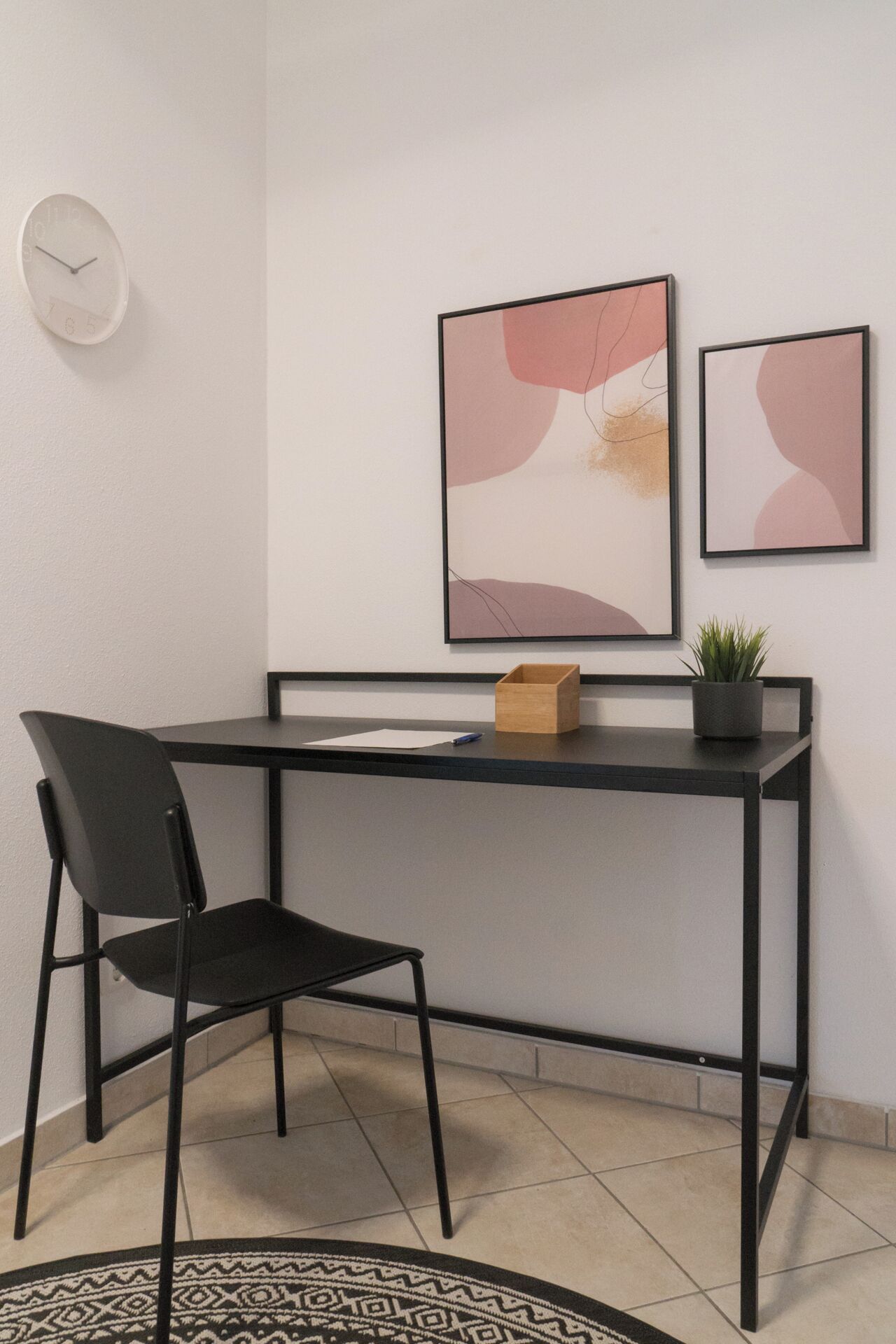 Un espacio de oficina moderno con un escritorio negro, una silla negra, un reloj y dos cuadros abstractos en la pared.