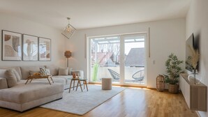 Un salon moderne avec un canapé, une table basse et une table à manger. Une grande fenêtre offre une vue sur les maisons à l’extérieur.