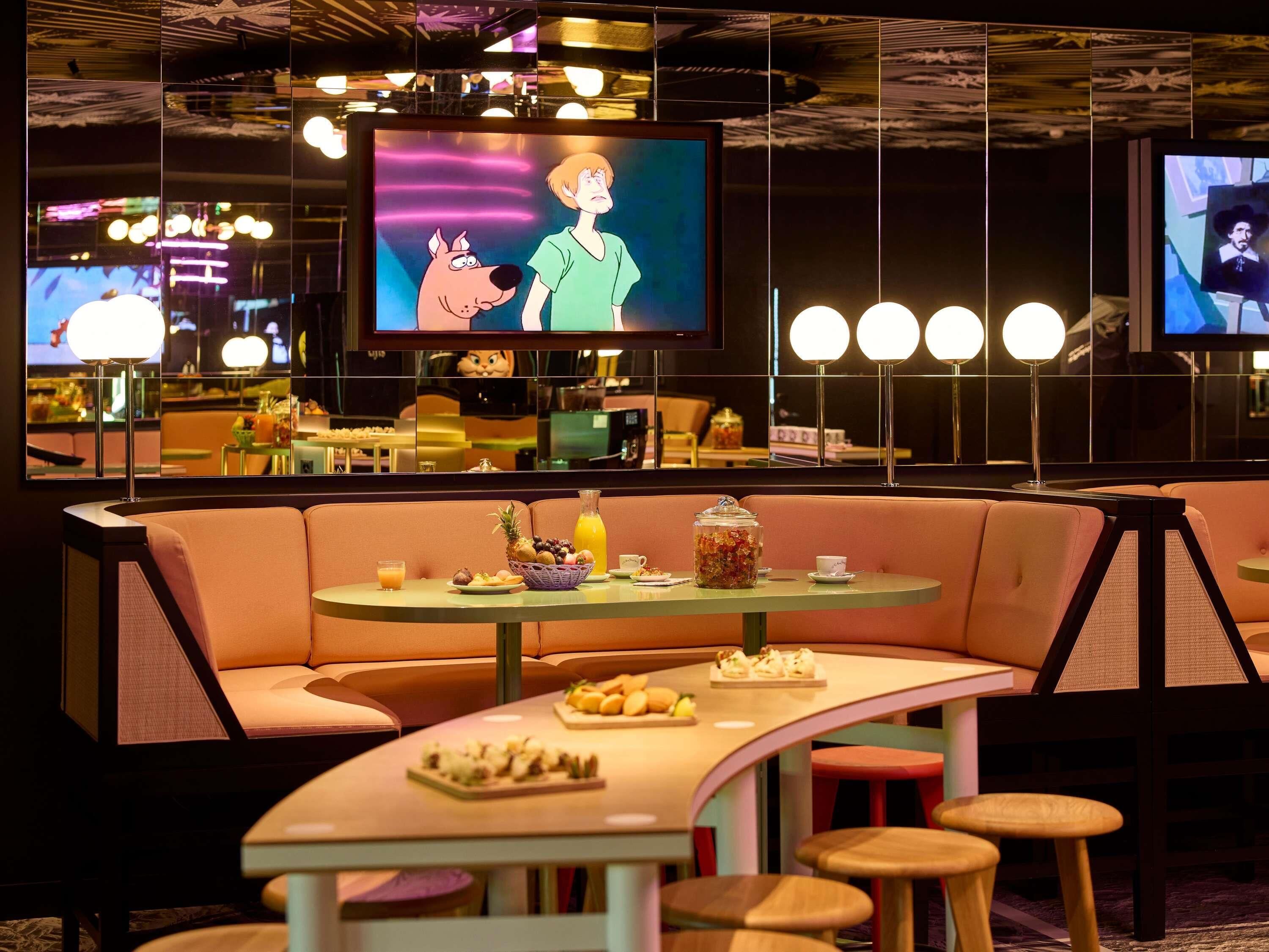 Un área de estar moderna con asientos, una mesa con comida y bebidas, y varias pantallas mostrando contenido.