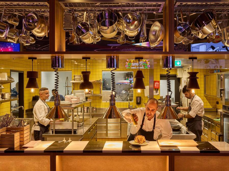 A kitchen with hanging pots and pans, chefs preparing food, and a yellow-tiled backsplash.