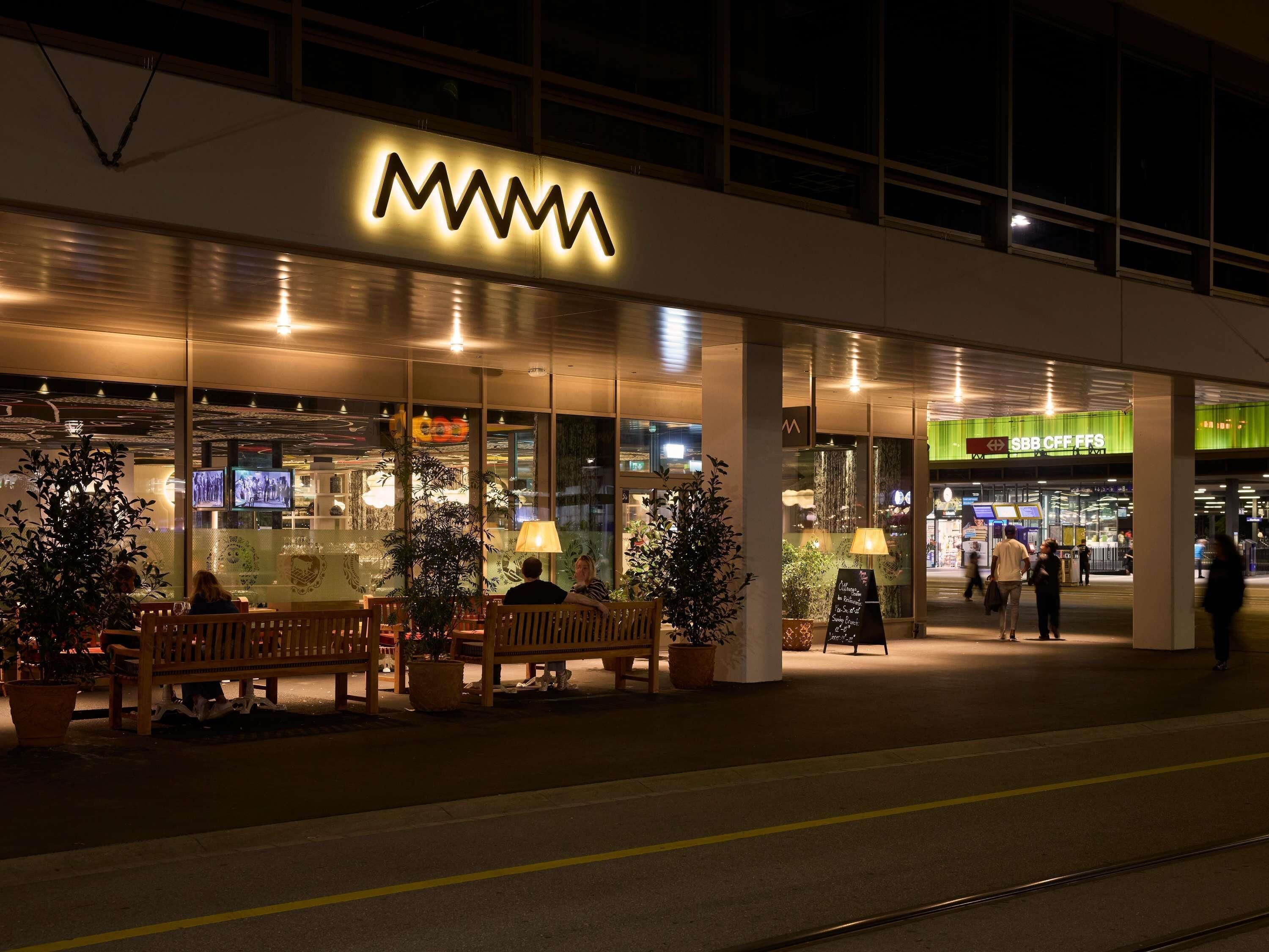A building with a prominent illuminated sign, outdoor seating with benches, and a green awning with signage.