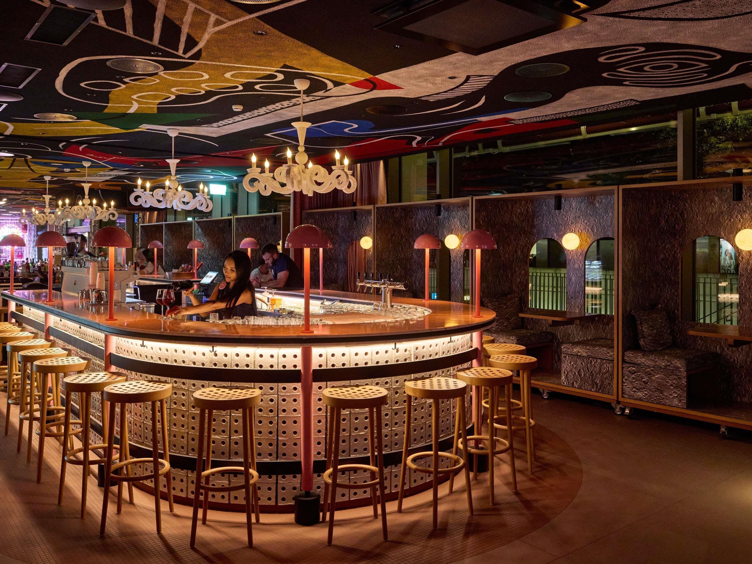 En bar med en cirkulær bardisk, flere barstole og et loft med abstrakte dekorationer.