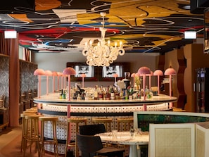 A bar area with a round table, stools, a chandelier, and a wall with a colorful abstract design. - Mama Shelter Zurich (Zurich)