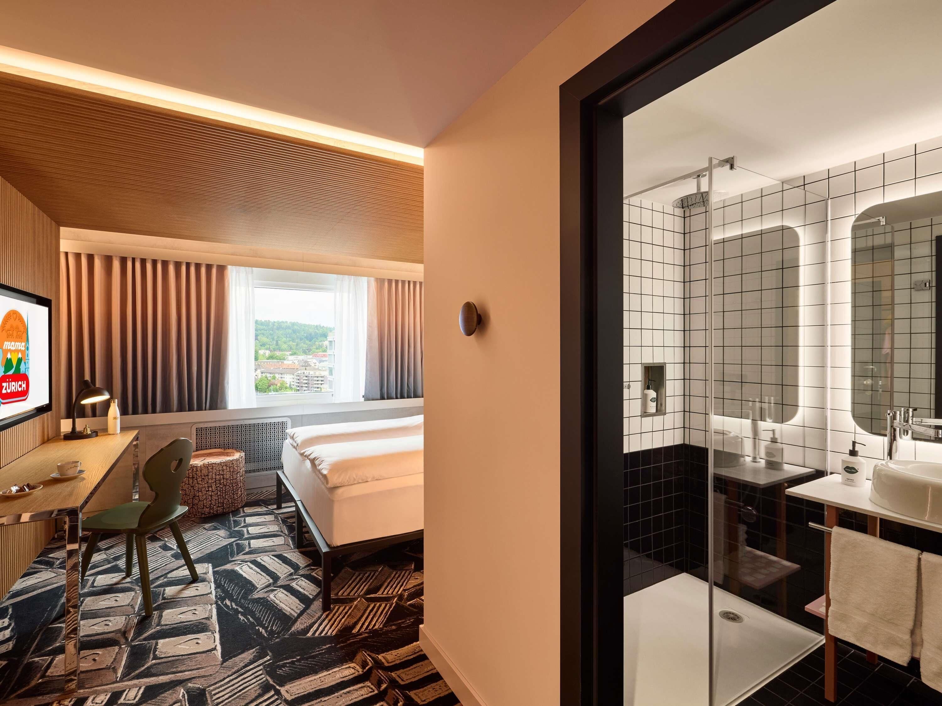 A hotel room with a bed, a desk with a chair, a window with curtains, and a bathroom with a mirror and sink.