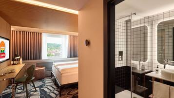 A hotel room with a bed, a desk with a chair, a window with curtains, and a bathroom with a mirror and sink.
