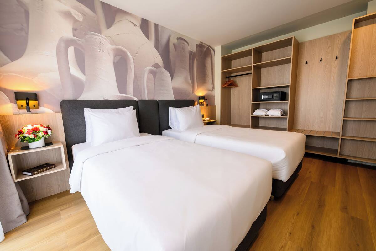 Une chambre d’hôtel avec deux lits, une armoire, une table de chevet avec une lampe et une étagère fixée au mur.