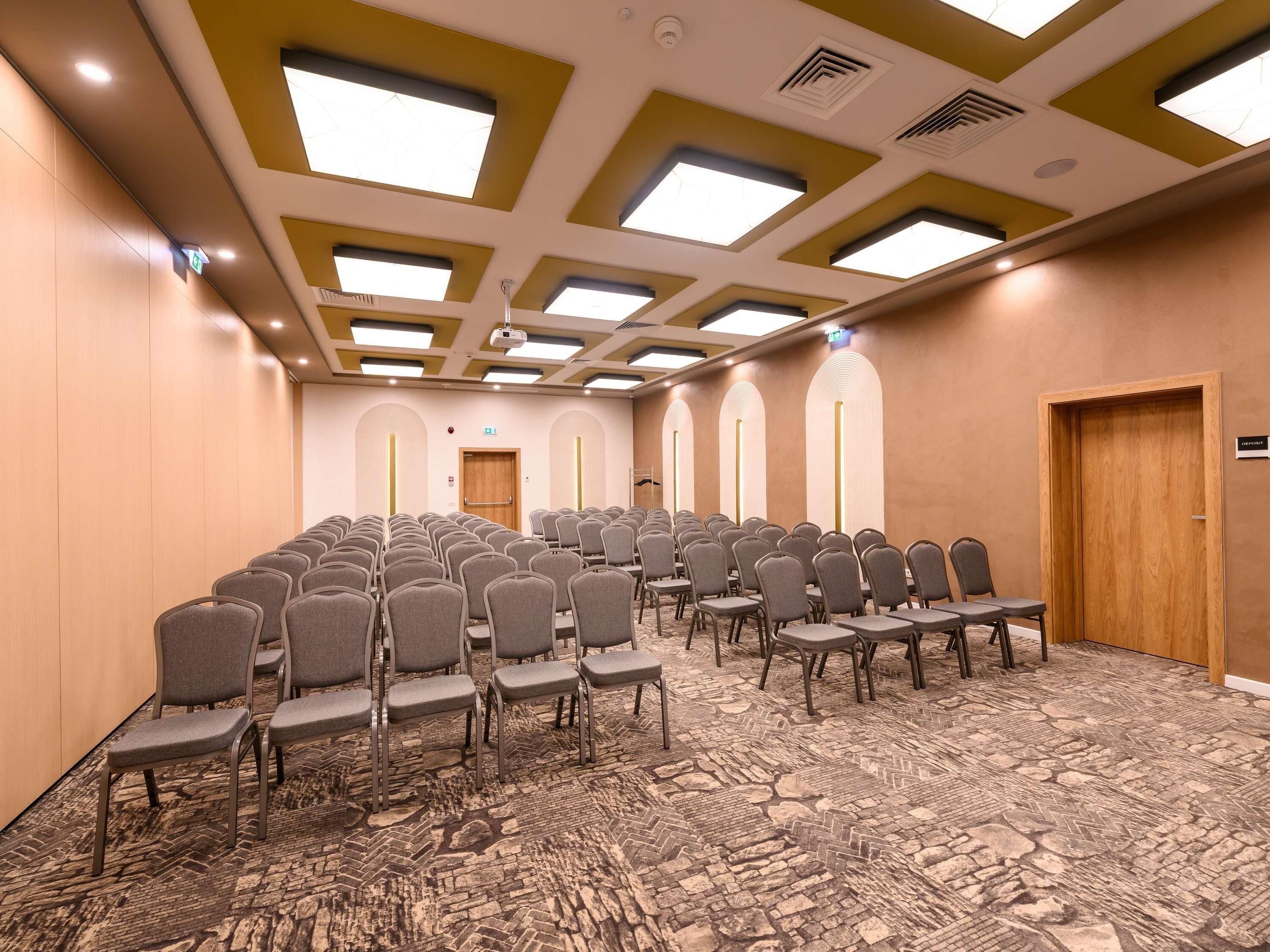 Sala de conferências com fileiras de cadeiras, carpete estampado e teto com iluminação embutida e claraboias.