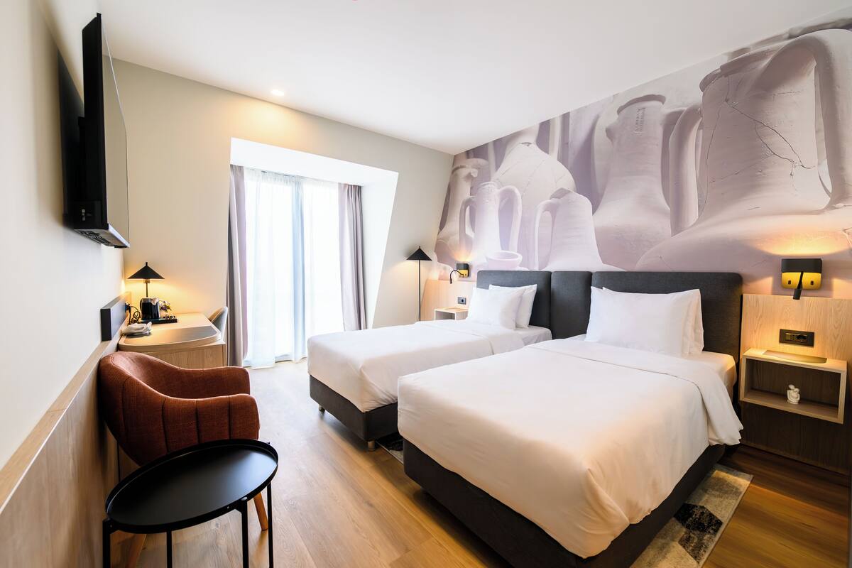 Une chambre d’hôtel avec deux lits, un téléviseur à écran plat fixé au mur, un bureau avec une chaise, une grande fenêtre avec des rideaux et une fresque murale.