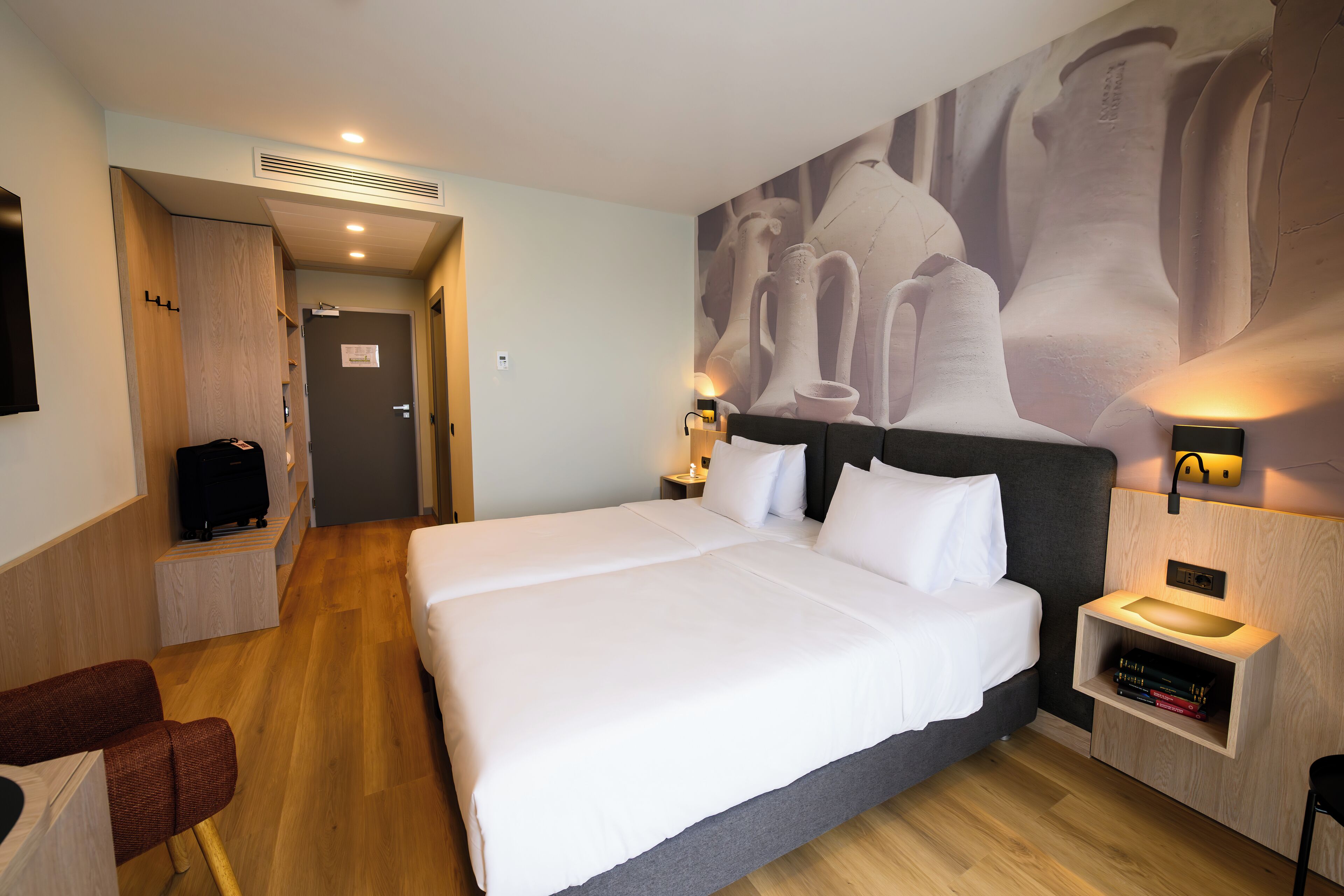 A hotel room with a large bed, a bedside table with a lamp, a wall-mounted shelf with books, a wooden wardrobe, and a television on a stand.
