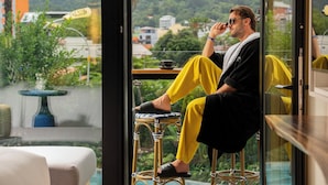 Una persona seduta su uno sgabello vicino a una finestra con vista su un panorama urbano e aree verdi.