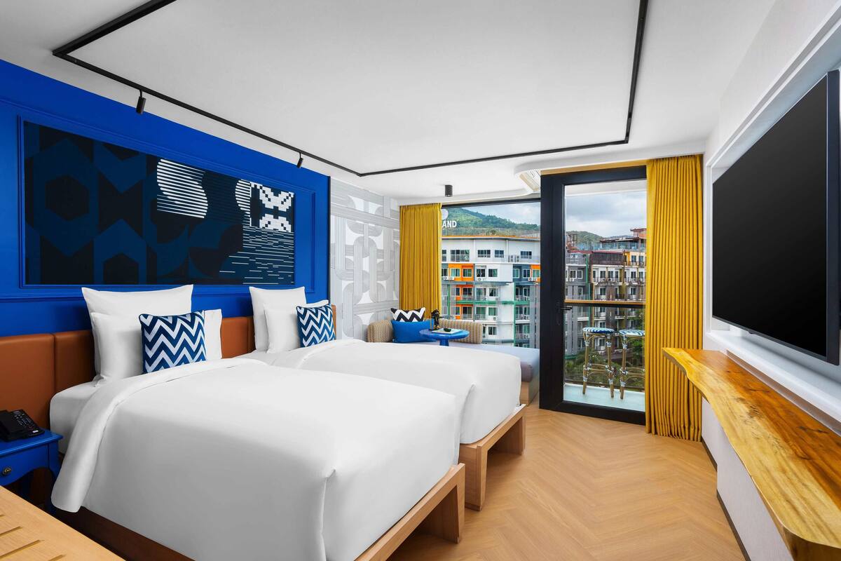 A modern hotel room with two beds, a large window with a view of buildings, a wooden desk, and a flat-screen TV.