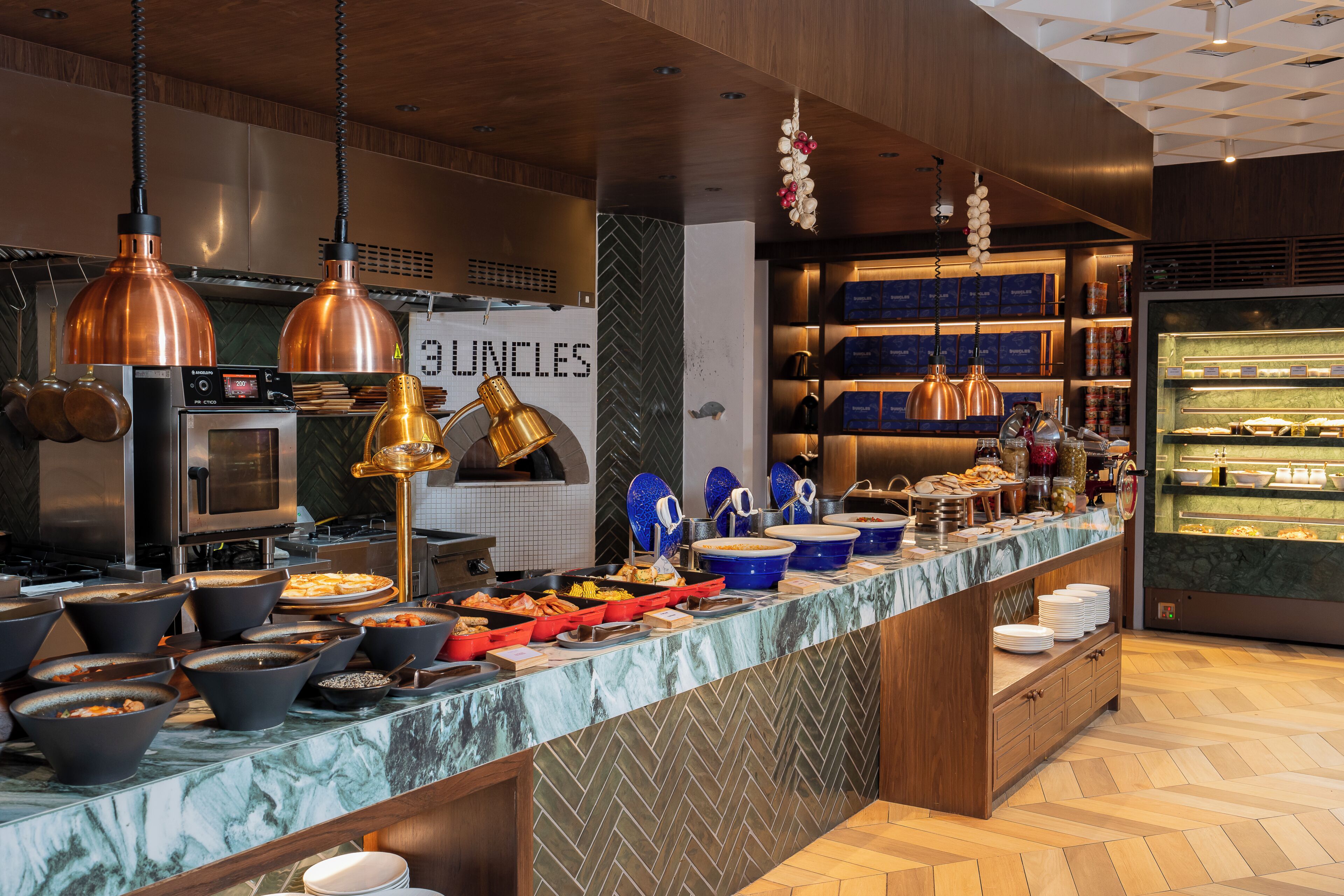 A buffet setup with various food items, a coffee machine, and a refrigerator in the background.